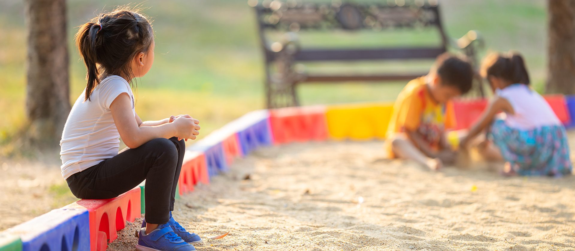 Little girl sitting lonely watching friends play at the playground